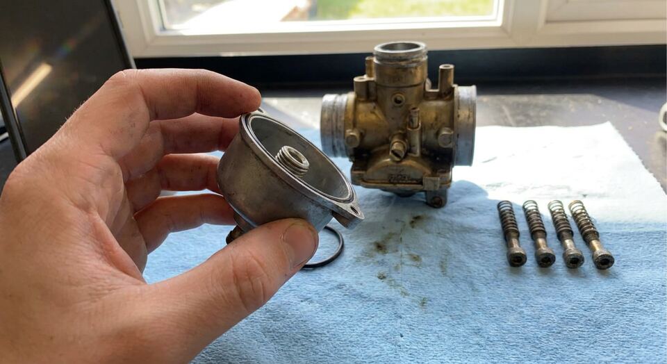 A hand holding the float bowl just unscrewed from the carburettor behind it, its four screws laid out on the rag